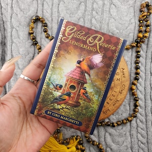May include: A deck of "Gilded Reverie Lenormand" cards held in a hand. The card box features a birdhouse scene with birds, gold accents, and the text "Gilded Reverie Lenormand" and "By Ciro Marchetti". The background includes a beaded necklace.