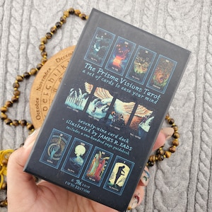 Op de afbeelding: Een zwart tarotkaartspel met de titel "The Prisma Visions Tarot" met illustraties en tekst. Het spel bevat 79 kaarten en een gids. De doos wordt vastgehouden in de buurt van een kralenarmband en een houten schijf met de maanden van het jaar.