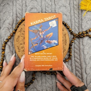 May include: A rectangular Karma Tarot deck box with an orange background and a central illustration of a winged figure holding a sword. The text reads Karma Tarot by Birgit Boline Erfurt, The 78-card Karma Tarot deck is a fascinating blend of color, motion and phantasmagoric art, and Complete With Instructions.