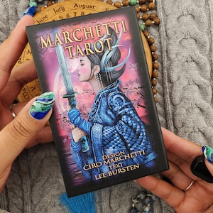 Può includere: Una scatola rettangolare per mazzo di tarocchi intitolata Marchetti Tarot con l'illustrazione di una persona in abiti blu a motivi geometrici che impugna una spada su un cielo rosa e viola. Il testo recita Design Ciro Marchetti e Text Lee Bursten.