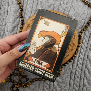 Puede incluir: Una caja de Tarot Aquarian vintage con una ilustración de una persona con sombrero y sosteniendo una flor. La caja es negra con detalles en naranja y crema. El texto "FOL" y "AQUARIAN TAROT DECK" son visibles.