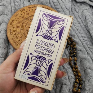May include: A vintage card game box with purple floral designs and the text "IL GIOCO DEI PERSONAGGI CARTE DA GIOCO". The box is held in a hand, with a wooden circular calendar and a beaded necklace in the background.