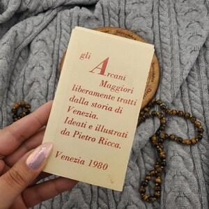 Puede incluir: Una tarjeta vintage con texto en italiano, sostenida contra una tela gris de punto trenzado. La tarjeta presenta el título "Arcani Maggiori" y el texto "Venezia 1980". También se ven un collar de cuentas y un disco de madera.