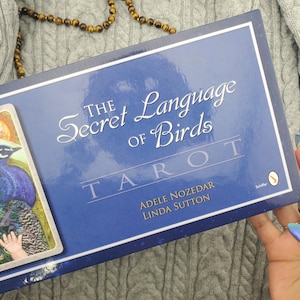 May include: A rectangular box for The Secret Language of Birds Tarot deck by Adele Nozedar and Linda Sutton. The box is dark blue with white text and features a partial illustration of a bird on the left side. The Schiffer logo is visible.