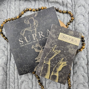 Peut inclure: Un livre noir intitulé Supra The Hidden Path of an Oracle et un étui de cartes noir assorti marqué Supra Oracle, tous deux ornés de dessins dorés de mains et de motifs célestes, disposés avec un collier de perles en œil de tigre.