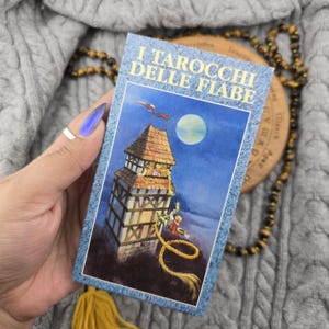 May include: A rectangular tarot card deck box titled I TAROCCHI DELLE FIABE in blue text. The illustration depicts a tall, timber-framed tower under a full moon, with a figure with long golden hair cascading from a window toward a person below.