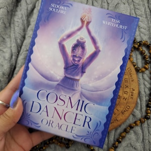 以下が含まれることがあります： セドナ・ソウルファイアとテス・ホワイトハーストによる「Cosmic Dancer Oracle」と題された長方形のオラクルカードデッキボックス。ボックスは紫と青のグラデーション背景で、両腕を頭上に上げて踊る人物のイラストが描かれています。