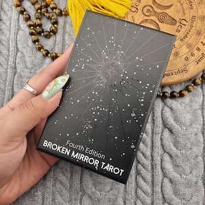 Puede incluir: Una caja de cartas de tarot negra con un diseño de estrella y el texto "BROKEN MIRROR TAROT Fourth Edition". La caja se sostiene en una mano con una uña pintada floral. Un disco de madera y un collar de cuentas están en el fondo.