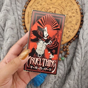 May include: A rectangular tarot card deck box featuring a red and black graphic illustration of a stylized figure with long red hair. The text CRUEL THING T A R O T is printed in white at the bottom of the box, which is framed by a pointed arch design.