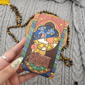 May include: A colorful, rectangular card with a stained-glass design featuring a night sky with stars, a crescent moon, and a candle. The card has a brown border and is held near a beaded necklace with a yellow tassel.