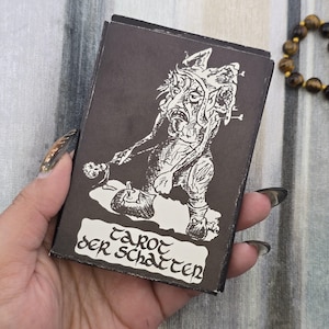 May include: A black tarot card box with a white illustration of a grotesque figure. The box has the words "Tarot der Schatten" in a white banner. The box is held in a hand with long, dark fingernails.