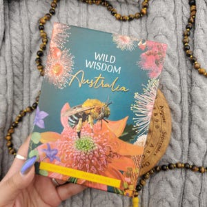 May include: A rectangular deck of cards titled Wild Wisdom Australia featuring a central illustration of a bee on a large orange flower. The box displays pink floral accents, green foliage, and the names Karin Roberts and Bonnie Hughes.