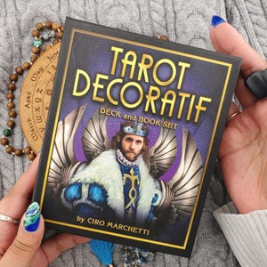 May include: A square Tarot Decoratif deck and book set box featuring a gold-lettered title and an illustration of a winged figure wearing a crown and fur-trimmed blue robe. The box includes the text by Ciro Marchetti on a purple textured background.