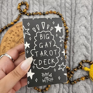 May include: A rectangular tarot deck box with a black background and white hand-drawn illustrations of stars and diamonds. The text reads BIG GAY TAROT DECK and Poppy the WITCH. The box is held against a backdrop of brown tiger eye beads.