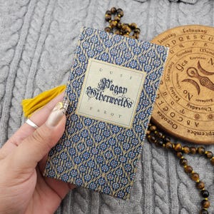 May include: A deck of "Pagan Otherworlds Tarot" cards with a blue and gold floral pattern. The card deck is held in a hand, with a yellow tassel and a wooden calendar wheel in the background. The text "Pagan Otherworlds Tarot" is visible.