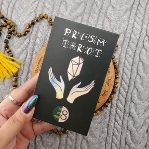 Op de afbeelding: Een zwarte Prism Tarot-kaartspeldoos met witte tekst en een holografisch kristalontwerp. De doos heeft de tekst "PRISM TAROT" en het logo "THE 8TH HOUSE". Het deck ligt op een houten onderzetter.