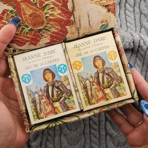 Op de afbeelding: Twee kaartspellen van Jeanne D'Arc in een decoratieve doos. Elk kaartspel heeft een illustratie van Jeanne d'Arc te paard, met tekst in het Frans. De doos heeft een bloemen- en landschapsontwerp.