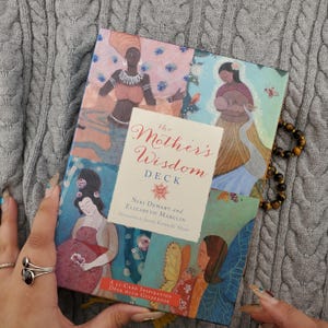 Puede incluir: El mazo Mother's Wisdom, un mazo de inspiración de 52 cartas con una guía, presenta ilustraciones coloridas de mujeres. La caja tiene una etiqueta color crema con el título y los nombres de los autores. Una pulsera de cuentas descansa cerca.