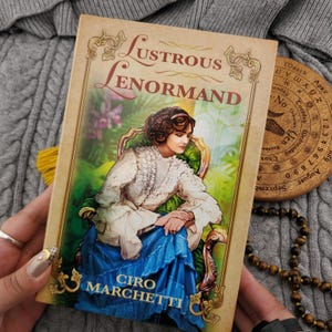 May include: A book titled "Lustrous Lenormand" by Ciro Marchetti. The cover features a vintage illustration of a woman in a blue dress seated in a chair. The book is held over a gray knitted surface, with a wooden divination tool visible.