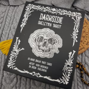 May include: A black tarot card box with the words "DARKSIDE SKELETON TAROT" in white. The box is decorated with skull and bone illustrations. The text "78 HAND DRAWN TAROT CARDS" is also visible. A yellow tassel and a wooden calendar are in the background.