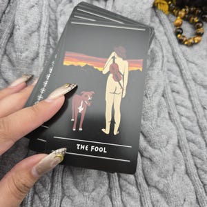 K&ouml;nnte beinhalten: Ein Tarotkartenspiel mit schwarzem Hintergrund. Eine Karte zeigt eine Illustration einer nackten Person mit einer Geige, einem Hund und dem Text "THE FOOL". Die Karten werden in einer Hand mit langen N&auml;geln gehalten.