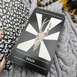 K&ouml;nnte beinhalten: Eine Tarotkarte mit schwarzem Hintergrund und wei&szlig;em geometrischen Design. Die Karte zeigt einen Hirschsch&auml;del mit floralen Akzenten und dem Wort "DEATH" unten. Die Karte ist Teil eines Decks.