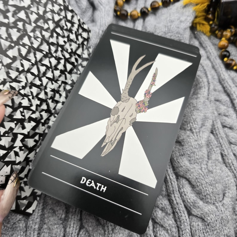 K&ouml;nnte beinhalten: Eine Tarotkarte mit schwarzem Hintergrund und wei&szlig;em geometrischen Design. Die Karte zeigt einen Hirschsch&auml;del mit floralen Akzenten und dem Wort "DEATH" unten. Die Karte ist Teil eines Decks.