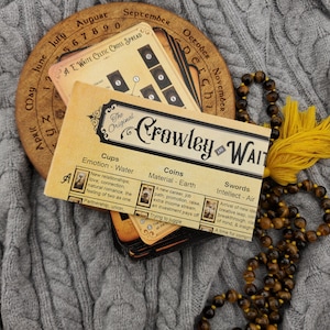 May include: A deck of Crowley and Waite tarot cards with a wooden calendar wheel and a beaded necklace. The cards have text and illustrations. The calendar wheel is brown with months and numbers. The necklace is brown and gold.