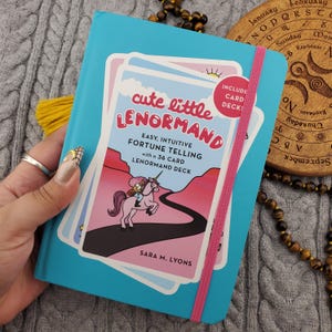 Puede incluir: Un mazo de cartas Lenormand azul turquesa con una banda elástica rosa. La portada presenta el texto "cute little LENORMAND" y una ilustración de una persona montando un unicornio. Incluye una baraja de 36 cartas.