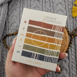 Może przedstawiać: Prostokątna talia kart Inner Compass Kids Cards w kremowym pudełku. Przód zdobią poziome paski w odcieniach brązu, pomarańczu, żółci, zieleni i błękitu oraz małe ikony, w tym odcisk łapy, liść, okulary i tęcza.