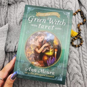 May include: The Green Witch Tarot deck box by Ann Moura with art by Kiri Østergaard Leonard. The green rectangular box features a circular illustration of a person pouring liquid into a cauldron, with white text and decorative border lines.