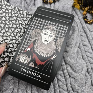 K&ouml;nnte beinhalten: Eine schwarze Tarotkarte mit dem Text "THE EMPEROR" unten. Die Karte zeigt eine Illustration einer Person mit Krone und R&uuml;schenkragen. Der Hintergrund hat ein sich wiederholendes Muster aus wei&szlig;en Kreuzen.