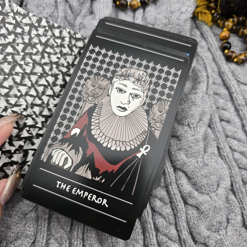 K&ouml;nnte beinhalten: Eine schwarze Tarotkarte mit dem Text "THE EMPEROR" unten. Die Karte zeigt eine Illustration einer Person mit Krone und R&uuml;schenkragen. Der Hintergrund hat ein sich wiederholendes Muster aus wei&szlig;en Kreuzen.