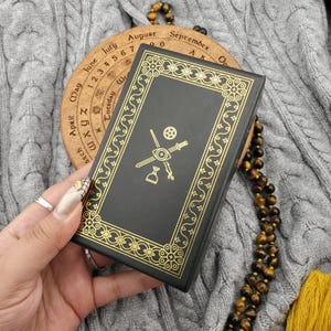 Könnte beinhalten: Ein schwarzes Buch mit goldenen Zierleisten und Symbolen, das vor einem hölzernen Kalenderrad gehalten wird. Der Einband des Buches weist einen goldenen Rand und Symbole auf. Eine Perlenkette und eine gelbe Quaste sind ebenfalls zu sehen.