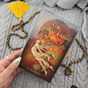 May include: A sealed deck of "The Gilded Tarot Royale" cards. The box features a woman in flowing fabric against a fiery background. The deck is held near a beaded necklace and a yellow tassel.