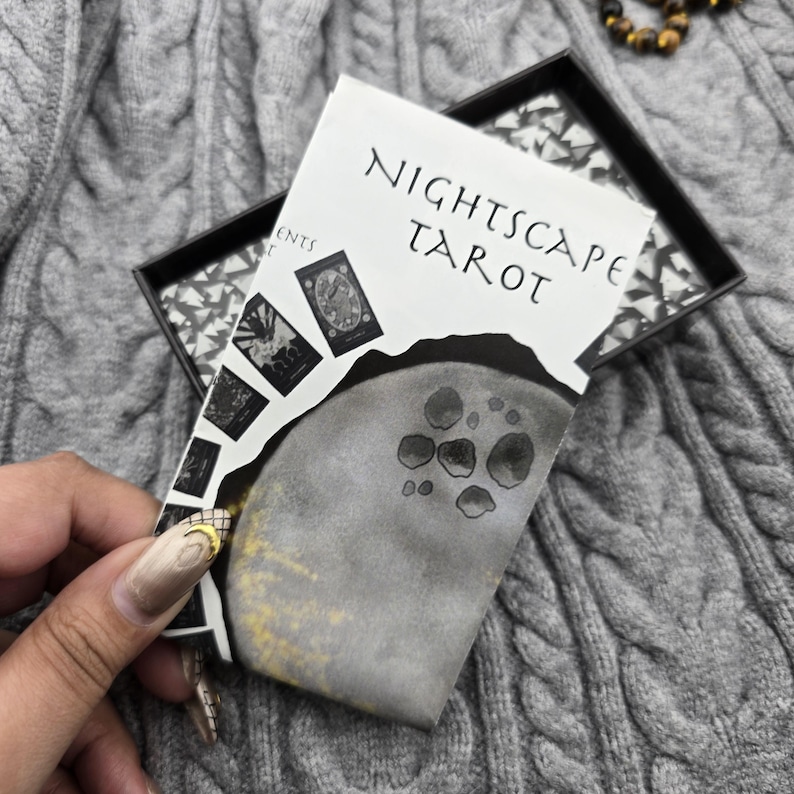 K&ouml;nnte beinhalten: Ein Tarotkartenspiel mit dem Titel "Nightscape Tarot" mit einem grau-wei&szlig;en Design. Das Deck zeigt Illustrationen des Mondes und anderer himmlischer Elemente. Die Karten werden in einer schwarzen Box mit gemustertem Innenraum auf einer grauen Strickoberfl&auml;che pr&auml;sentiert.