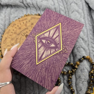 May include: A rectangular deck of cards with a deep purple cover featuring a central gold diamond frame enclosing a detailed eye illustration. The design includes radiating lines and small gold dots, creating a sunburst effect across the surface.