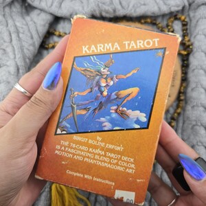 May include: The Karma Tarot deck box features an orange background with the title in white text. A central illustration depicts a winged figure holding a sword against a blue sky. Text below identifies the author Birgit Boline Erfurt and describes the 78-card deck as a blend of colour, motion, and phantasmagoric art, including instructions.