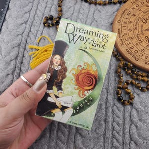 May include: A deck of "Dreaming Way Tarot" cards by Rome Choi. The box features an illustration of a woman in a black dress and top hat, with a sun and crescent moon design. The text "TORAH" is visible. U.S. GAMES SYSTEMS, INC. is printed at the bottom.
