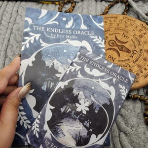 Puede incluir: Se muestran dos copias de "The Endless Oracle" de Eric Maille, un juego de adivinación. Las portadas presentan una ilustración en azul oscuro y blanco de una escena de bosque. También se ven un tablero de adivinación de madera y un collar de cuentas.