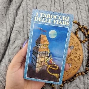 May include: A rectangular tarot card titled I TAROCCHI DELLE FIABE featuring a blue-toned illustration of a tall tower under a full moon. A figure with long golden hair hangs from the window, while another figure climbs the hair below.
