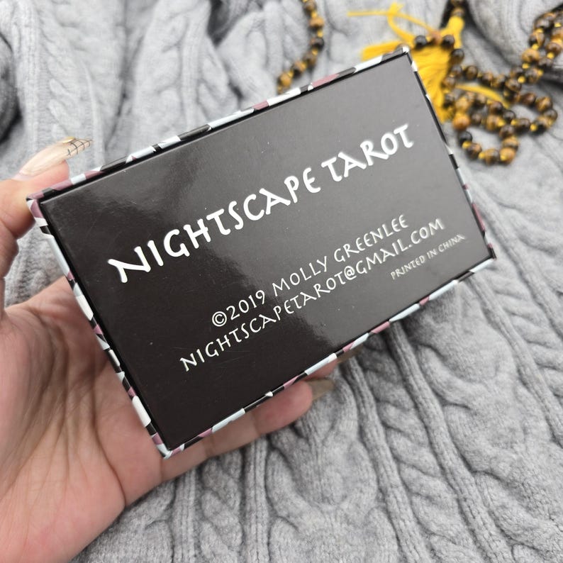 K&ouml;nnte beinhalten: Eine schwarze Tarotkartenbox mit den wei&szlig;en Worten "NIGHTSCAPE TAROT". Die Box enth&auml;lt auch die Copyright-Informationen und die E-Mail-Adresse. Die Box wird vor einem grauen Strickhintergrund gehalten.