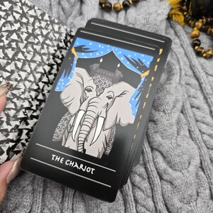 K&ouml;nnte beinhalten: Ein Tarotkartenspiel mit einem grauen Elefanten mit wei&szlig;en Sto&szlig;z&auml;hnen und einem dekorativen Kopfschmuck, vor einem blauen Vorhanghintergrund. Die Karte tr&auml;gt die Aufschrift "THE CHARIOT" in Wei&szlig; auf schwarzem Grund. Das Deck liegt auf einer grauen Zopfstrickoberfl&auml;che.