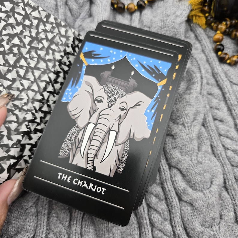 K&ouml;nnte beinhalten: Ein Tarotkartenspiel mit einem grauen Elefanten mit wei&szlig;en Sto&szlig;z&auml;hnen und einem dekorativen Kopfschmuck, vor einem blauen Vorhanghintergrund. Die Karte tr&auml;gt die Aufschrift "THE CHARIOT" in Wei&szlig; auf schwarzem Grund. Das Deck liegt auf einer grauen Zopfstrickoberfl&auml;che.