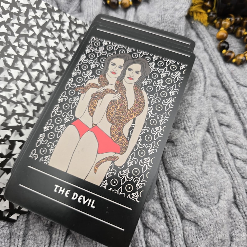 K&ouml;nnte beinhalten: Eine schwarze Tarotkarte mit den Worten "THE DEVIL" am unteren Rand. Die Karte zeigt eine Illustration von zwei Frauen mit einer Schlange, die sich um sie wickelt. Die Frauen tragen rote Unterteile.