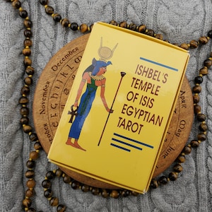 Könnte beinhalten: Eine gelbe Tarotkartenbox mit dem Text "ISHBEL'S TEMPLE OF ISIS EGYPTIAN TAROT" und einer Illustration einer ägyptischen Figur. Die Box befindet sich auf einer Holzscheibe mit eingravierten Monaten und einer Perlenkette.