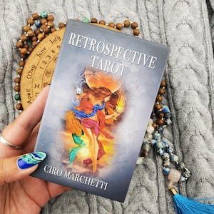 Può includere: Una scatola rettangolare per mazzo di tarocchi intitolata Retrospective Tarot di Ciro Marchetti. La copertina presenta un'illustrazione centrale di una figura drappeggiata in tessuti colorati e fluenti che regge due calici dorati davanti a una grande luna arancione.