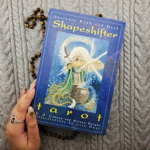 May include: A blue tarot card box with the title "Shapeshifter" in yellow lettering. The box includes a book and deck, with illustrations of a woman and an eagle. The word "tarot" is at the bottom. A beaded necklace is next to the box.