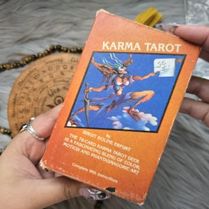 Könnte beinhalten: Ein orangefarbenes Karma-Tarotkartenspiel mit einer blauen Illustration einer mythischen Figur, die ein Schwert hält. Der Text "Karma Tarot" steht oben, mit "Complete With Instructions" unten.