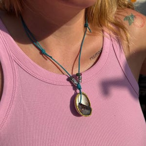 Può includere: Una collana con un cordino color verde acqua, due perline cilindriche color argento e un piccolo ciondolo a forma di conchiglia irregolare. La conchiglia ha l'interno bianco, il centro marrone scuro e il bordo dorato, fissata da un nodo.